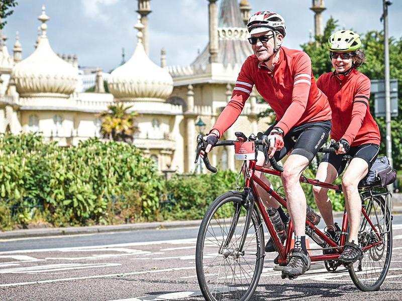 Two people riding on a tandem. They are cycling past Brighton Pavillion.