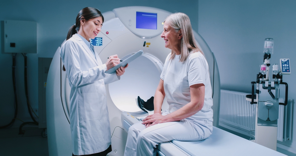Woman sat on table of CT scan talking to a doctor who's holding a  clipboard.
