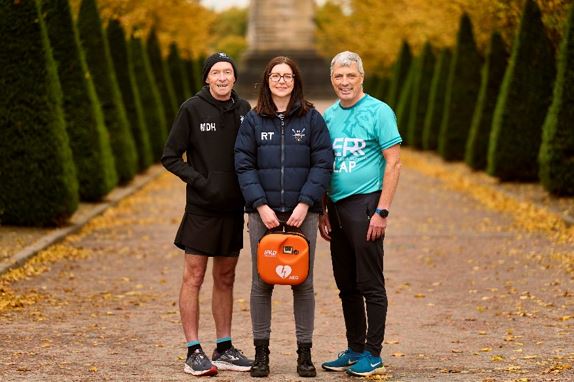 Two men are stood on either side of a woman who is holding a defibrillator