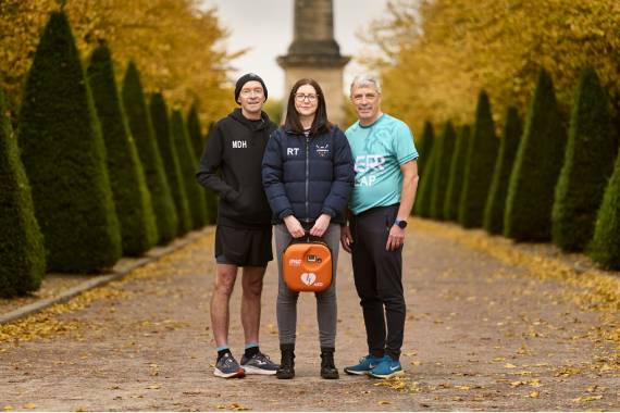 Two men are stood on either side of a woman who is holding a defibrillator