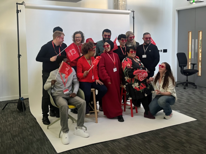Group of colleagues taking part in a workplace photoshoot, posing with British Heart Foundation flags and props in front of a studio backdrop.