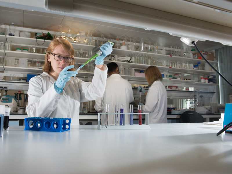 Researcher using a pipette to transfer liquid into a test tube in a laboratory, with other researchers working in the background.