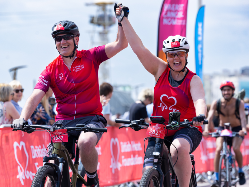 Two participants in a British Heart Foundation fundraising cycle ride raise their joined hands while crossing the finish area on bicycles.