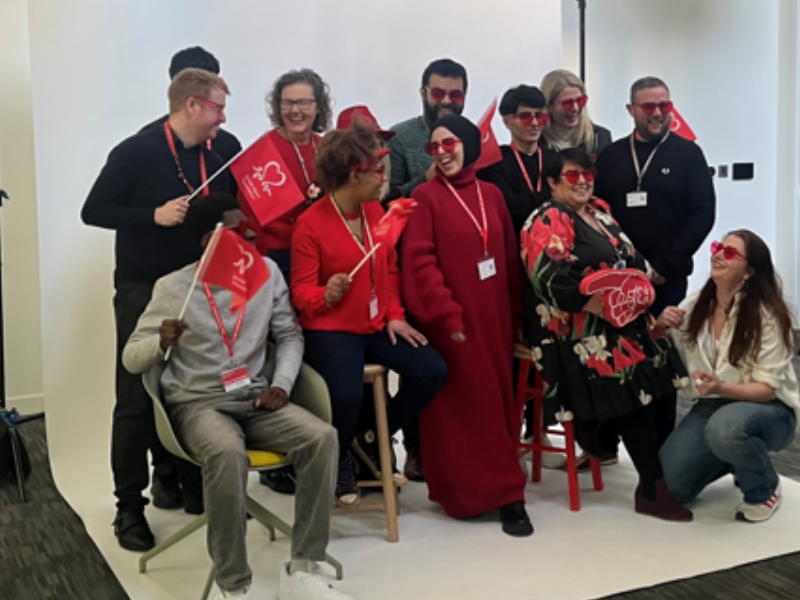 Group of BHF staff and volunteers posing together at an indoor event. Several people are holding BHF-branded props, including flags and signs, while wearing red glasses and event lanyards.