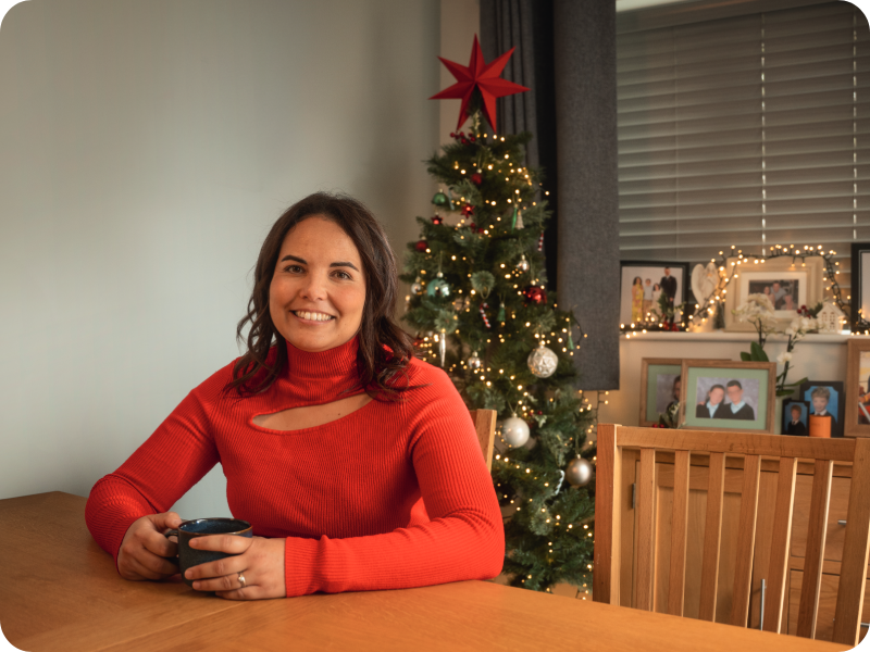 Carly sits at a wooden table holding a mug, with a Christmas tree and framed photos in the background.