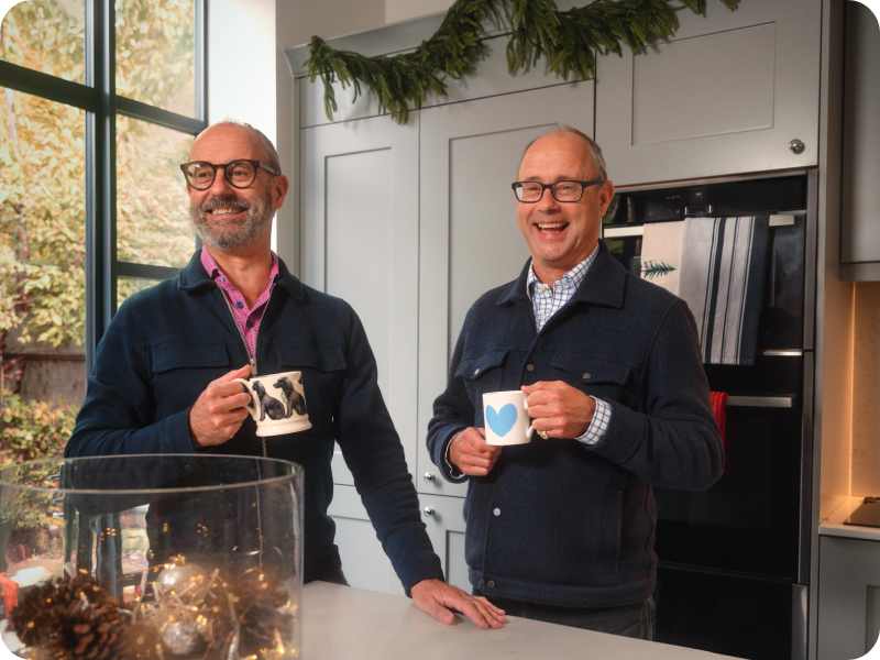 Patrick and Peter stand holding mugs in a kitchen, laughing and smiling, a festive decoration in the foreground.