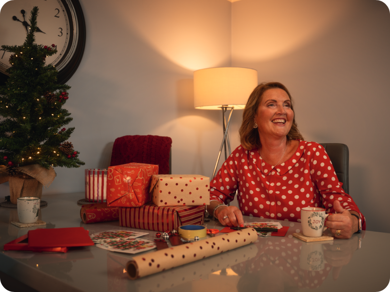 Rebecca sits at a table with presents and wrapping paper, and a small Christmas tree.