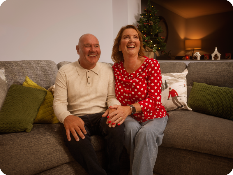 Rebecca sits on a brown sofa next to her husband in front of a Christmas tree, they both laugh.