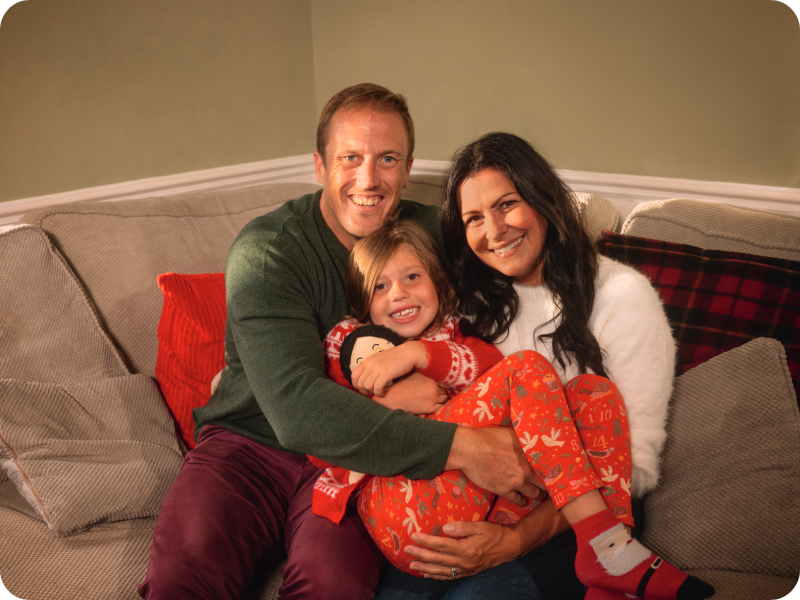 Amalie on the sofa hugging a doll with her parents' arms around her, wearing red Christmas pyjamas and festive socks.