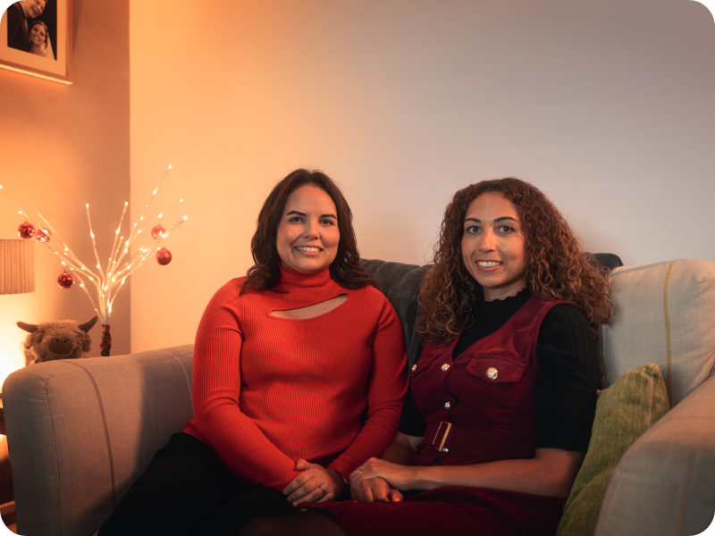 Carly and her sister Jodie on a sofa in a warmly-lit room.