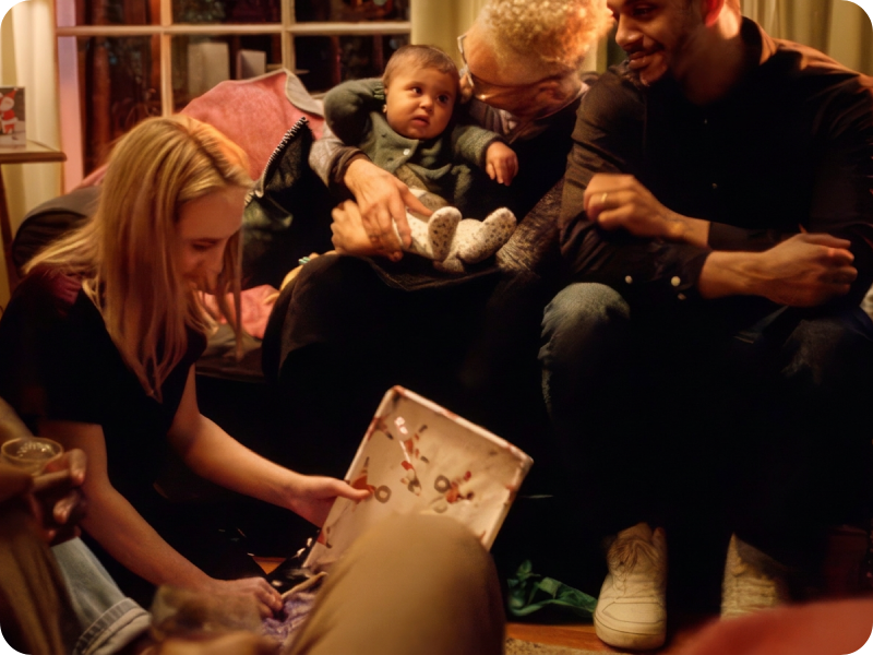 A few people sit around a person opening Christmas presents, one of them holds a baby.