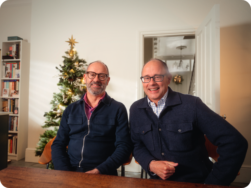 Patrick and Peter sit at a table smiling, wearing navy jackets, with a Christmas tree behind them.