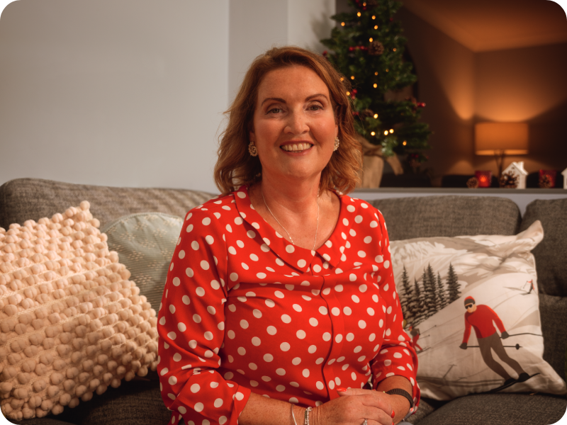 Rebecca sits on a sofa in front of a Christmas tree smiling, wearing a red spotty blouse.