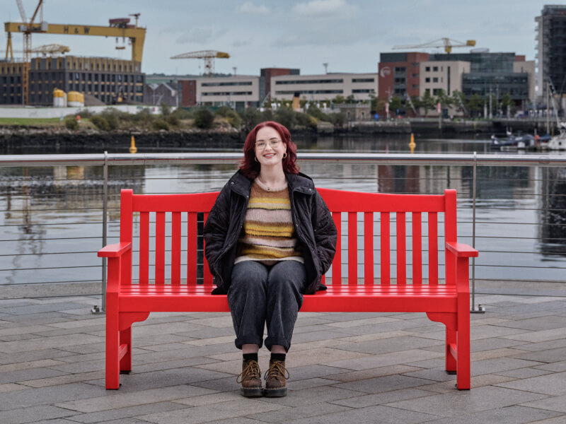 Caoimhe, sitting on a red bench near a river.