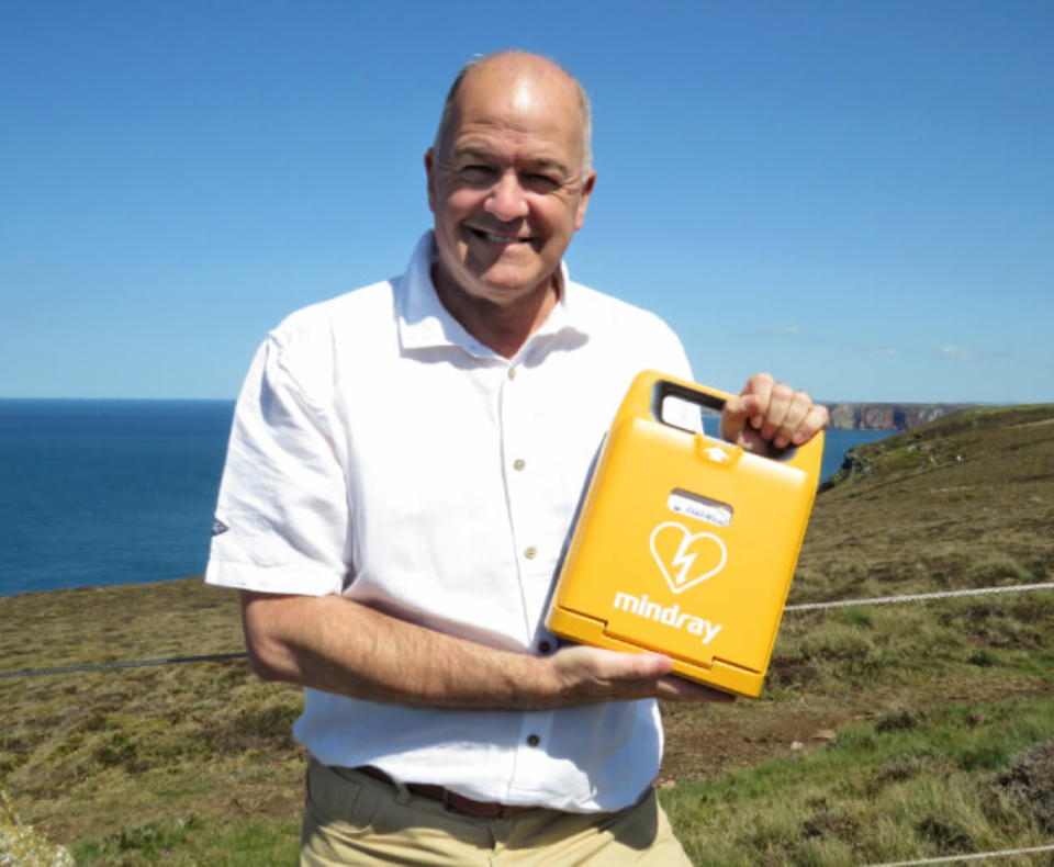 A photograph of Paul holding a defibrillator on a clifftop in St Agnes.