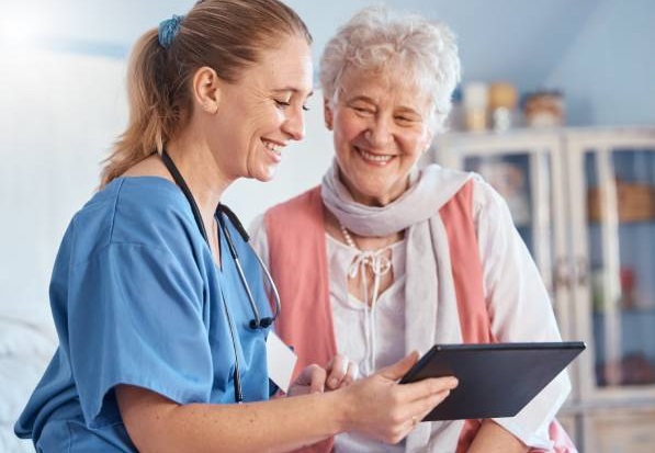 a female nurse with blonde hair sitting next to an older female patient and smiling as she shows the patient her tablet computer