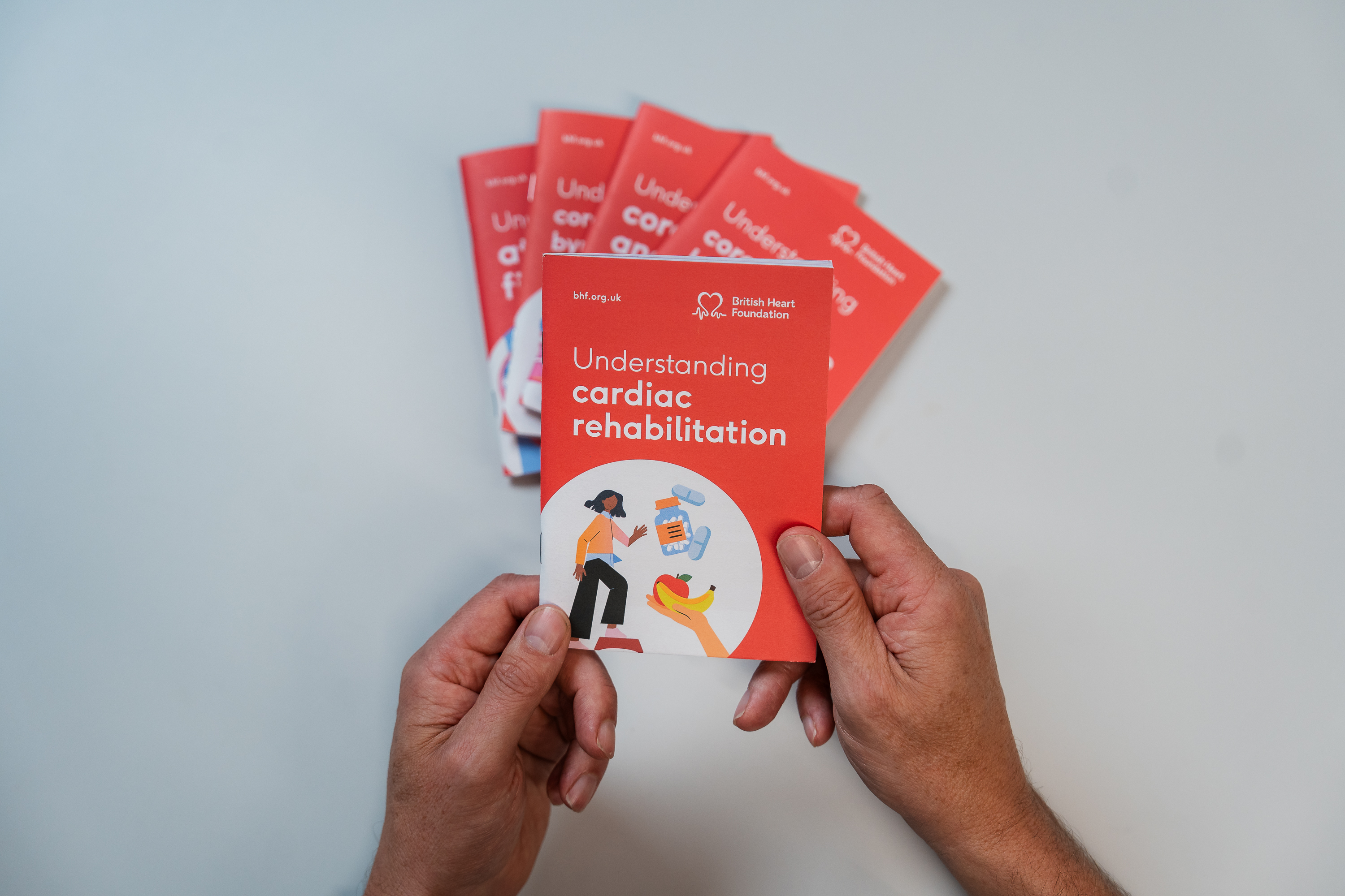 Persons hands holding a small red booklet titled Understanding cardiac rehabilitation. A white background with blurred out other small red booklets behind.