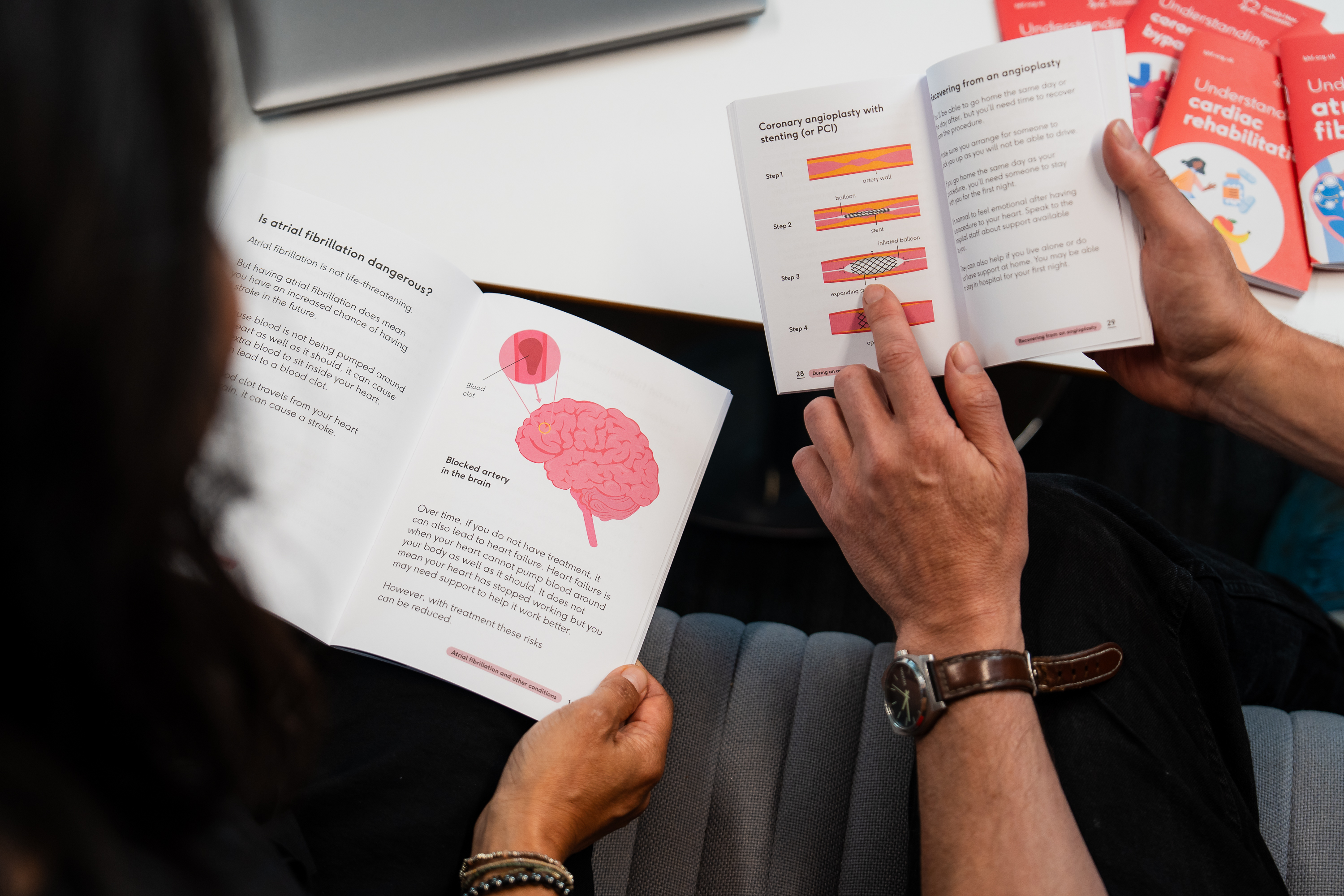 Someone with long dark hair holding a booklet open and reading the information, next to another person holding a booklet. You can mostly see their hands and arms holding the two booklets with a table in the background.