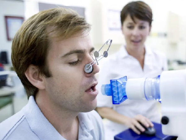 a male patient with blonde hair breathing into the mouthpiece of a medical device while a female nurse with brown hair stands behind him