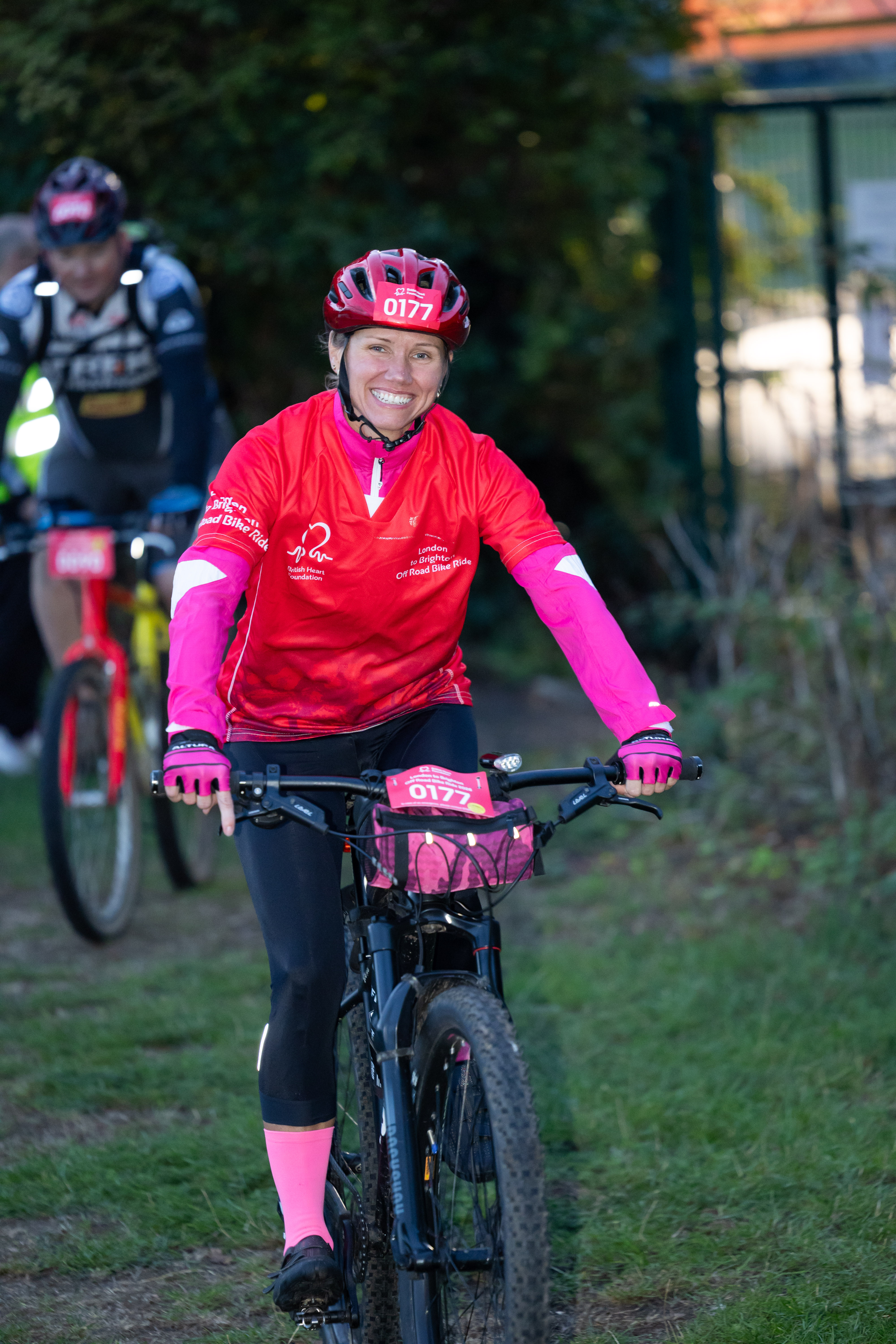 Female cyclist in a red BHF top and helmet riding a mountain bike along a wooded trail, with another cyclist behind.
