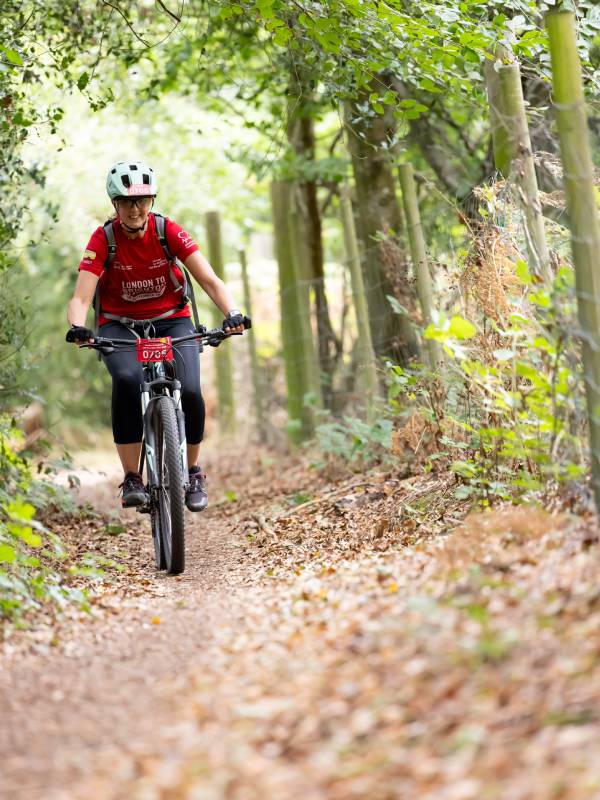 A man in a red BHF top and helmet riding a mountain bike along a narrow wooded trail.