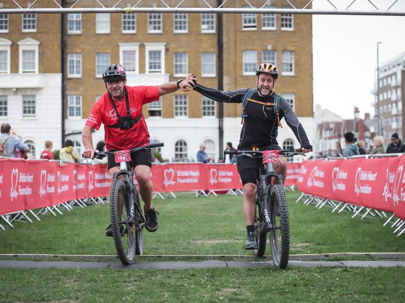 Two male cyclists riding mountain bikes through the finish area of a BHF cycling event, reaching across to touch hands.