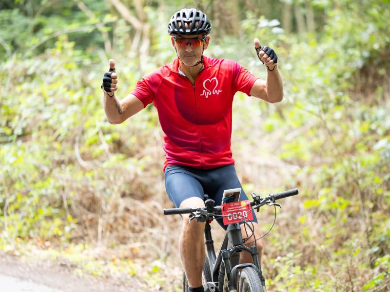 Cyclist in a red British Heart Foundation top riding a mountain bike along a wooded trail, with both thumbs raised.