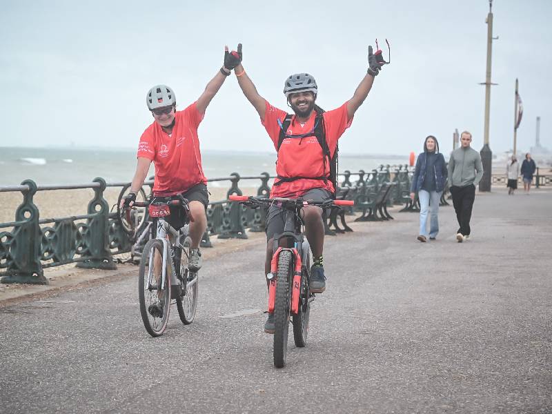 Two cyclists in red tops riding along a seafront promenade, raising their arms as people walk nearby.