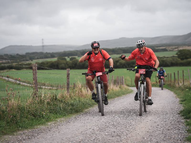 Three cyclists in red tops riding mountain bikes along a gravel track through the countryside.