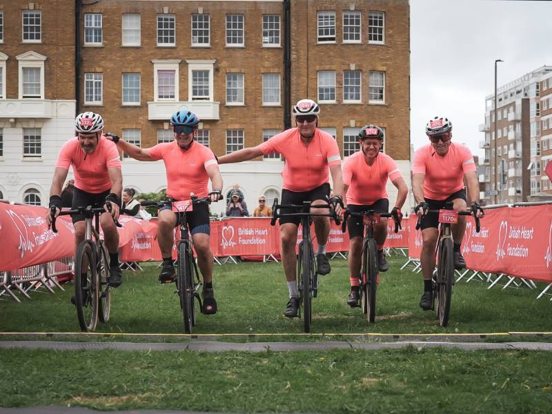 Five cyclists in matching red tops riding mountain bikes across a grassy finish area at a BHF cycling event.