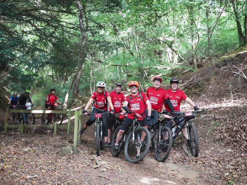 Group of cyclists in red tops with mountain bikes, gathered together on a wooded trail.