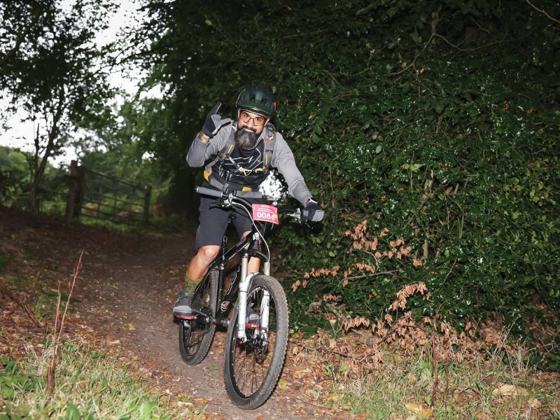 Cyclist wearing a helmet riding a mountain bike along a wooded trail, with one hand raised.
