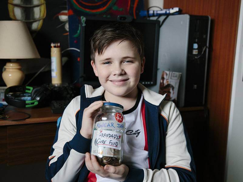 Boy holding up a jar with money with a label that says 'Grandad's cure fund'.