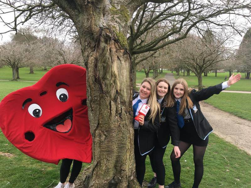 Hearty the BHF mascott peeking out from behind a tree with 3 school girls in school uniforms posing on the other side of the tree.