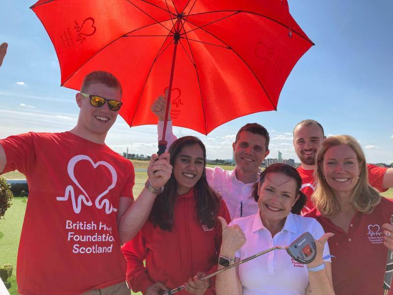 Group of 6 people smiling with one holding a golf club and another holding up an umbrella to cover them from the sun