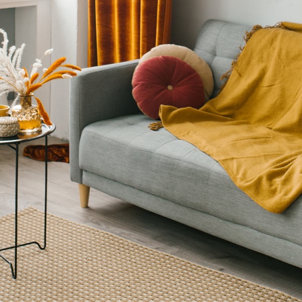 An autumnal living room with a grey sofa, draped in a yellow ochre throw. A table with orange flowers can be seen beside it.