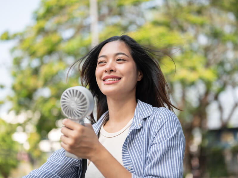 A woman holding a fan to her face while outdoors in the sunshine.