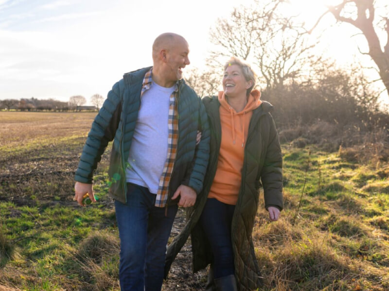 A man and a woman wearing green jackets walk in a green field while smiling at each other