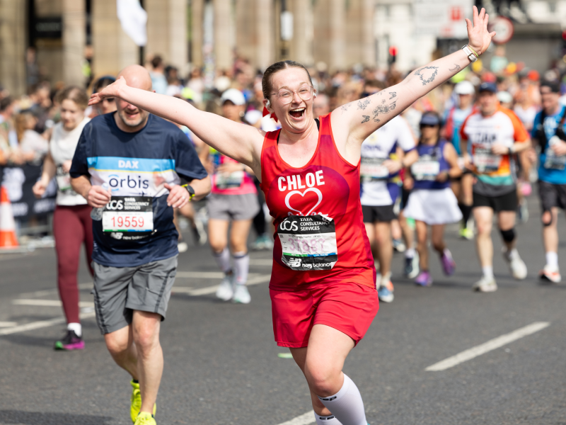 Runner in a red BHF outfit stretches both arms out while taking part in a marathon, surrounded by other participants on a city street.