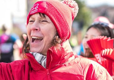 an older woman wearing a red bobbled hat and a red jacket smiling as she cheers on volunteers at an event
