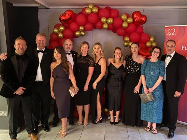 a group of men and women in smart eveningwear stood in a line and smiling in front of an arch made from red and gold balloons