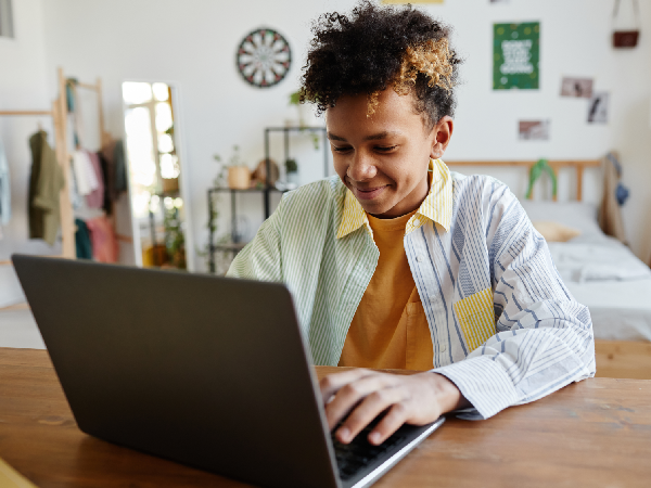 a black teenage boy sitting at a desk in his bedroom and smiling as he types on his laptop