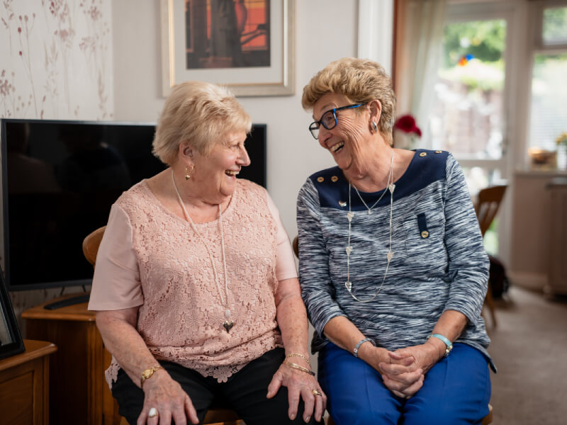 Shirley and Maureen sitting in a living room smiling at each other