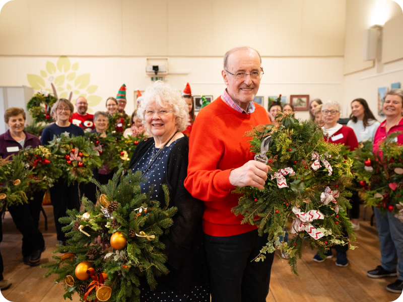 People holding handmade Christmas wreaths at a festive community workshop, with two participants standing in the centre displaying decorated wreaths.