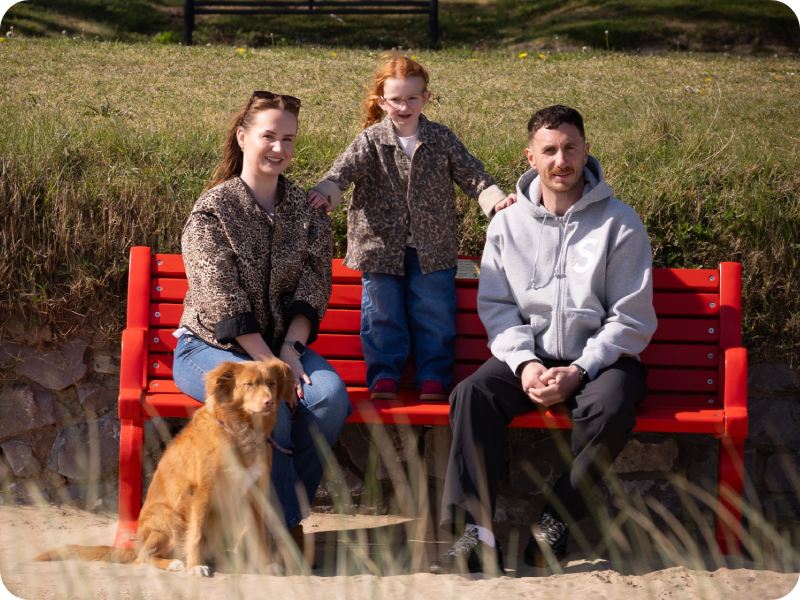 The Mariani family sits on a red BHF bench by the beach with a dog, outdoors on a sunny day.
