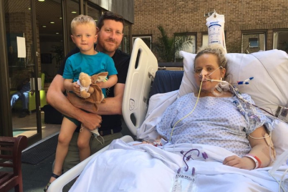 Woman lying in hospital bed hooked up to medical equipment with husband and young son standing next to her