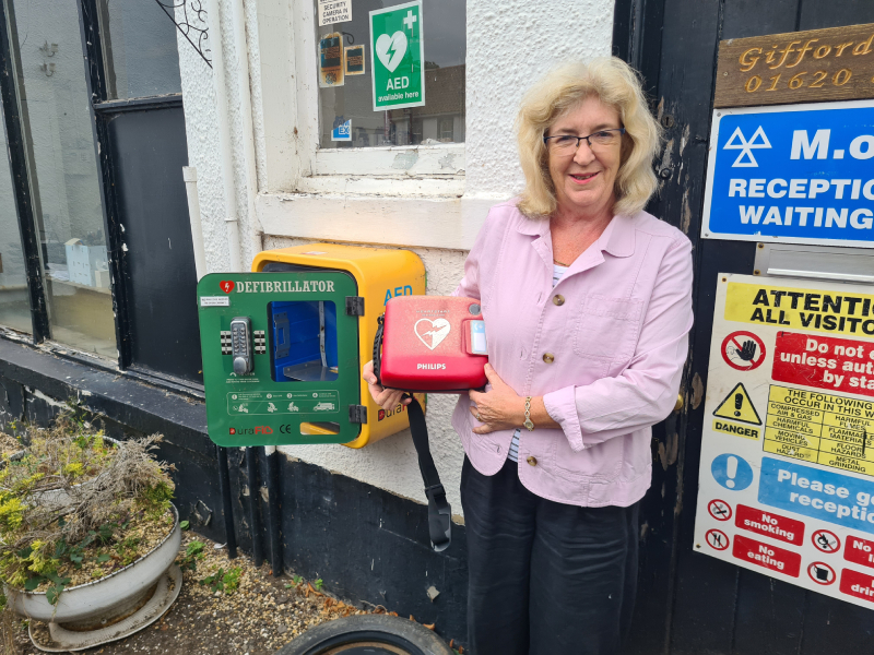 Eleanor stands outside a building and holds a red defibrillator