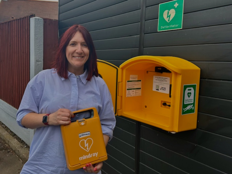 Lianne stands outside of a building holding a yellow defibrillator