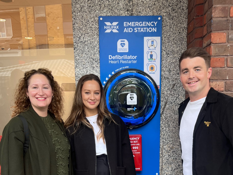 Two women and a man stand outside of a building smiling next to a defibrillator