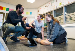 Two students practising CPR on a mannequin whilst a teacher is crouched next to them observing.